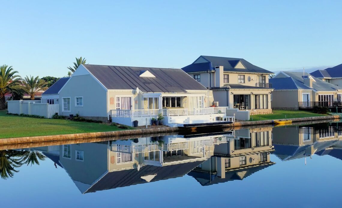 Luxury modern homes reflecting on a peaceful, clear lake during daylight, showcasing architectural beauty.
