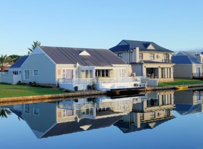 Luxury modern homes reflecting on a peaceful, clear lake during daylight, showcasing architectural beauty.